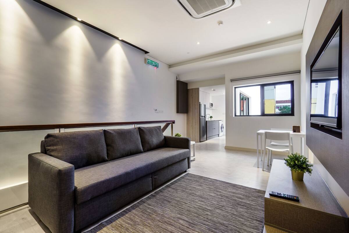 A modern living room with a grey sofa, a small table with a plant, a TV mounted on the wall, and a kitchen area in the background.
