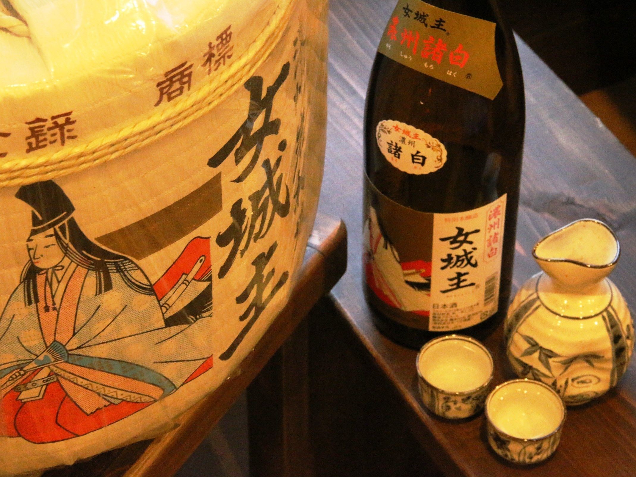 Una botella tradicional de sake japonés con etiqueta, un recipiente de cerámica grande y tres pequeñas tazas de cerámica sobre una superficie de madera.