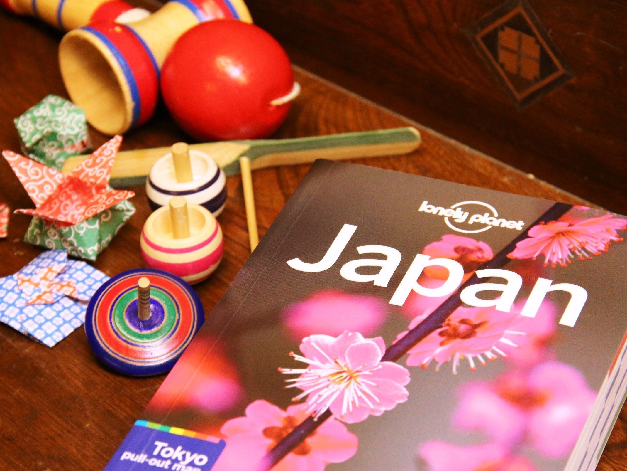 Ein Buch mit dem Titel 'Lonely Planet Japan' mit floralem Design, auf einer Holztischfläche zusammen mit verschiedenen bunten Kreiselspinnen und einer kleinen Karte mit dem Aufdruck 'Tokyo Ausklappkarte'.