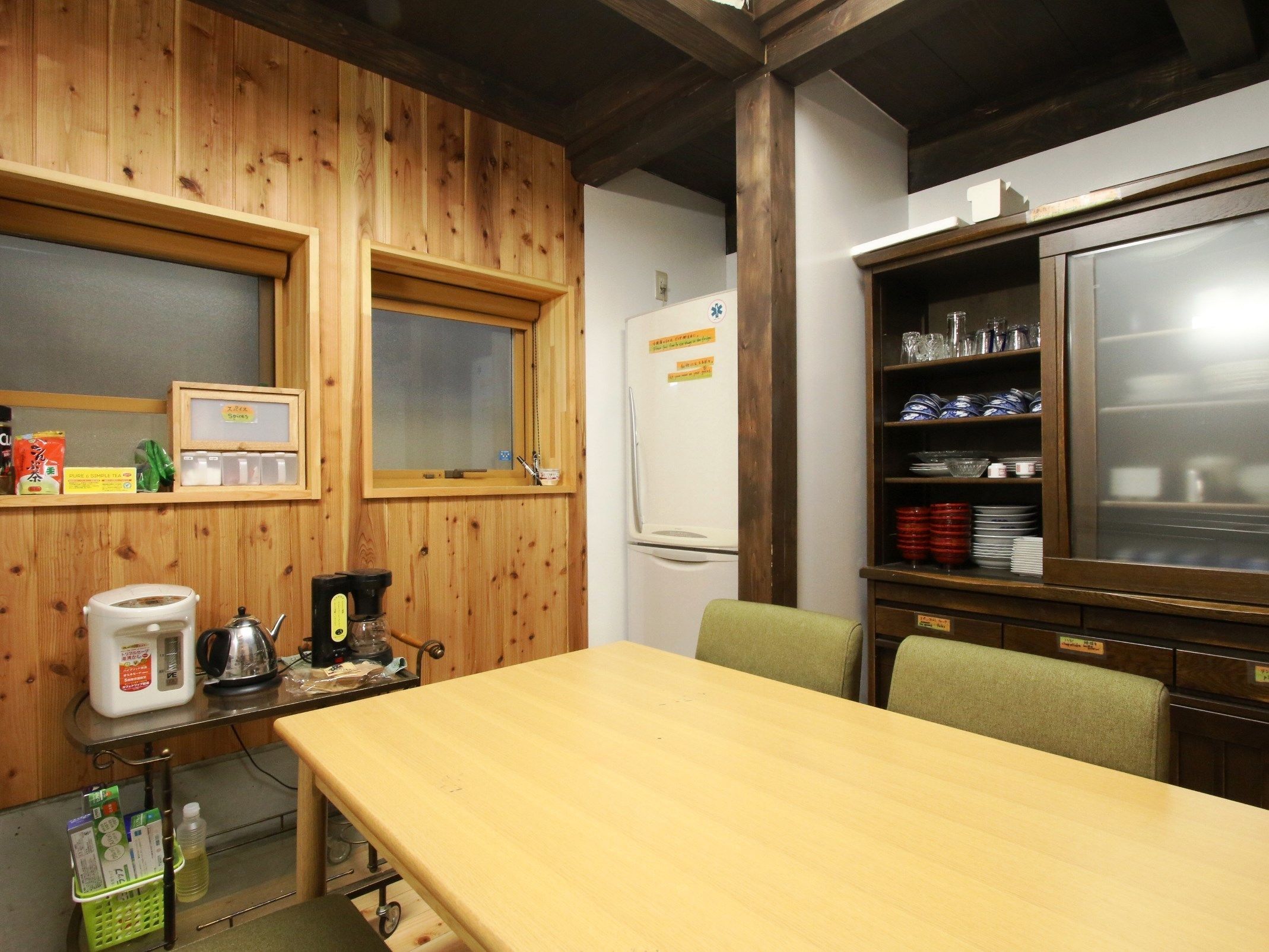 A wooden-paneled room with a wooden table, chairs, a refrigerator, a shelf with dishes, and a window.