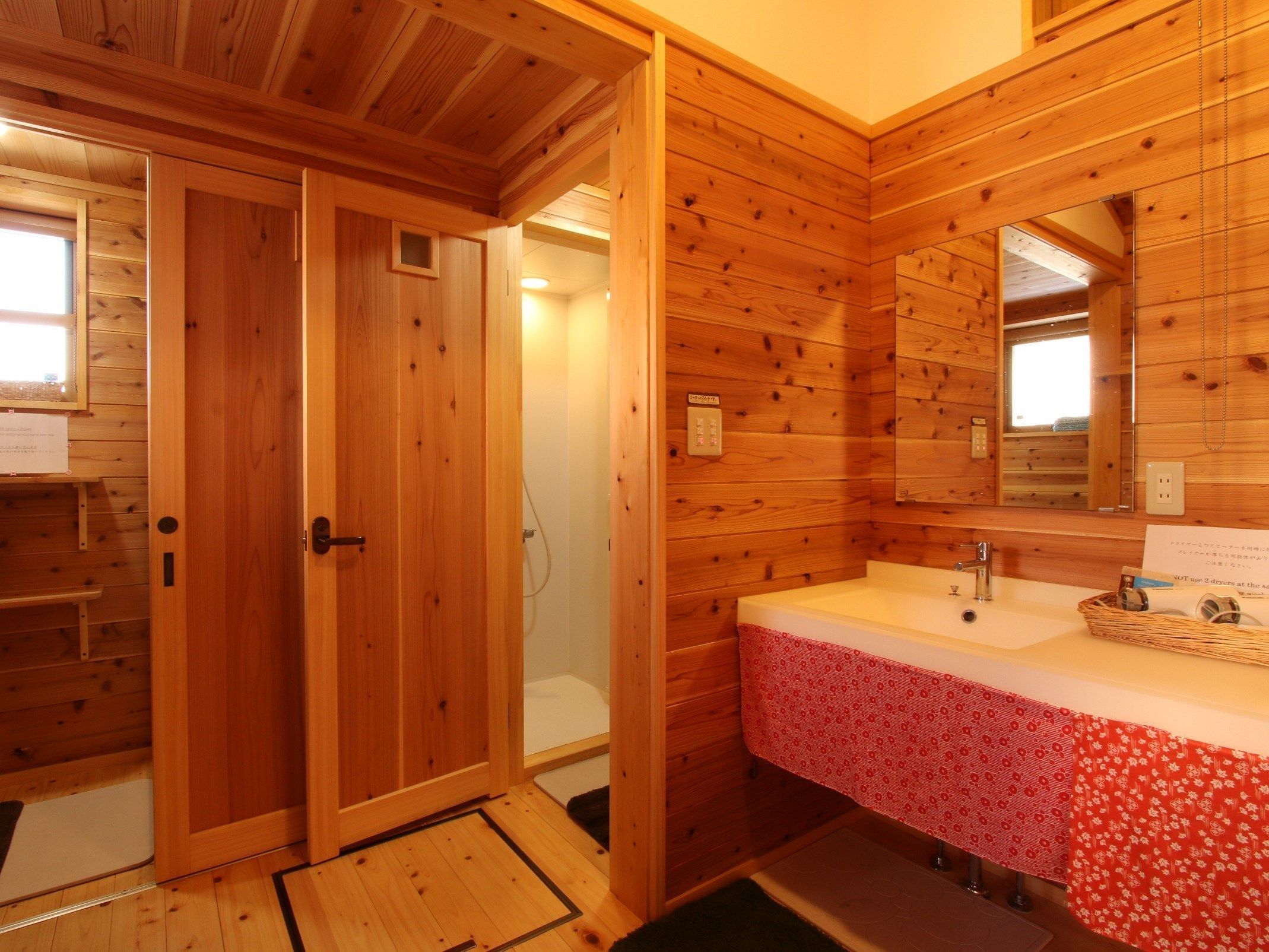 Een houten badkamer met een houten deur, een douche, een wastafel met een rode handdoek met een patroon, en een spiegel.