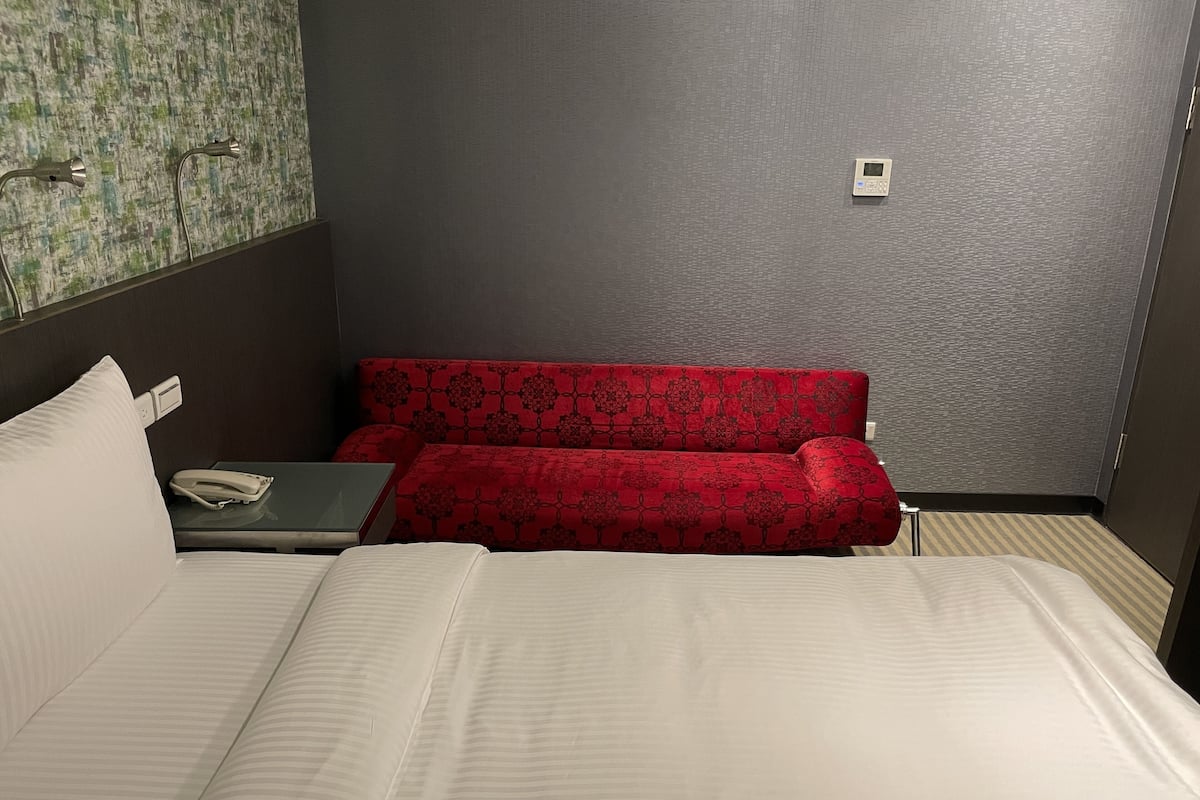 A hotel room with a bed, a red sofa, a small table with a phone, and a thermostat on the wall.