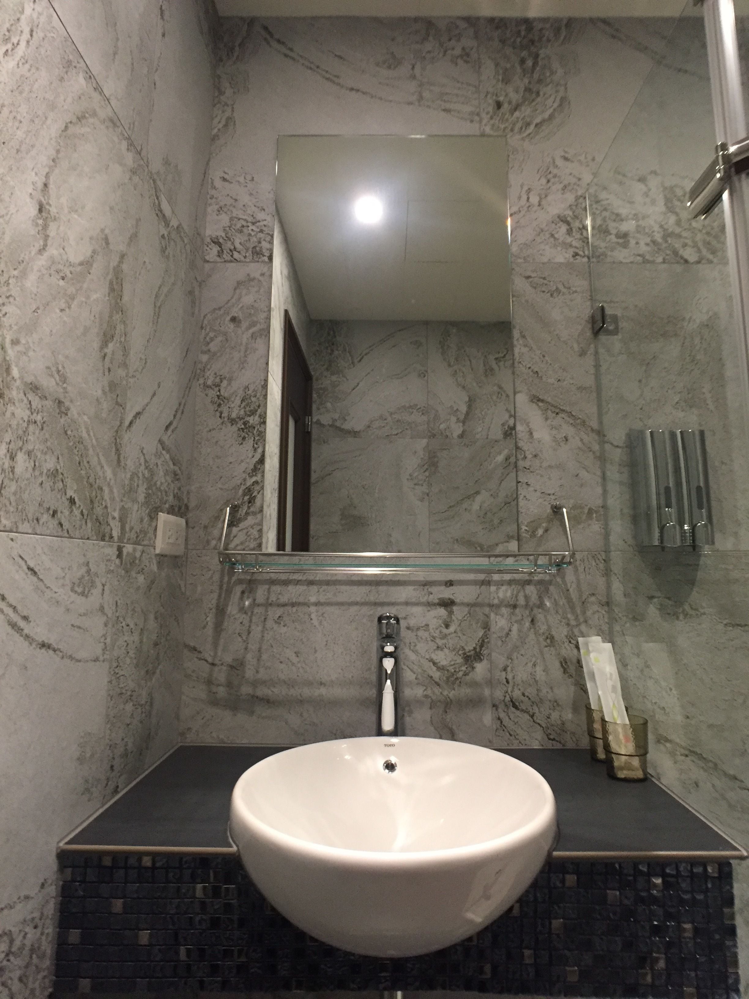 A bathroom with a marble wall, a white sink, and a glass shelf above it.