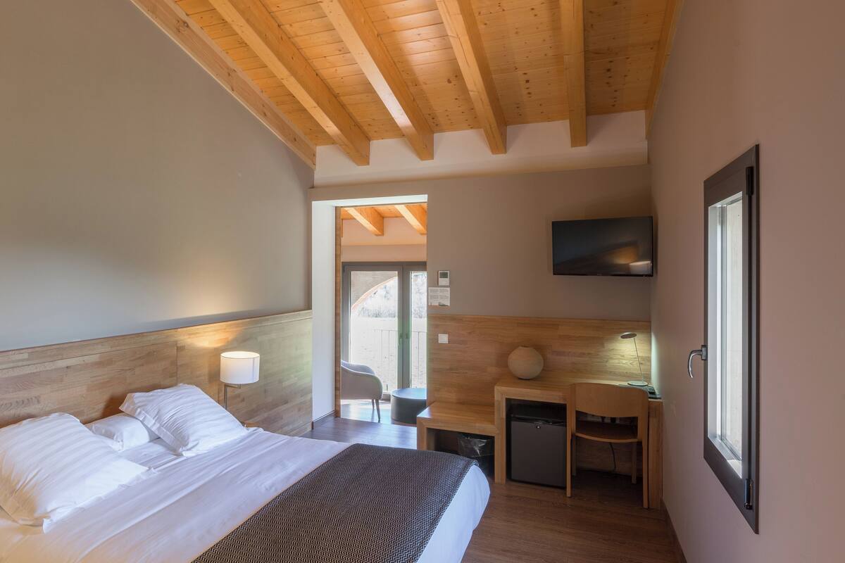 A bedroom with a wooden headboard, a bed with white linens, a desk with a chair, a small refrigerator, and a television mounted on the wall.