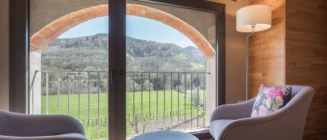 A room with a large arched glass door leading to a balcony with a view of a green landscape and hills. There is a round table and two chairs with a colorful pillow on one.