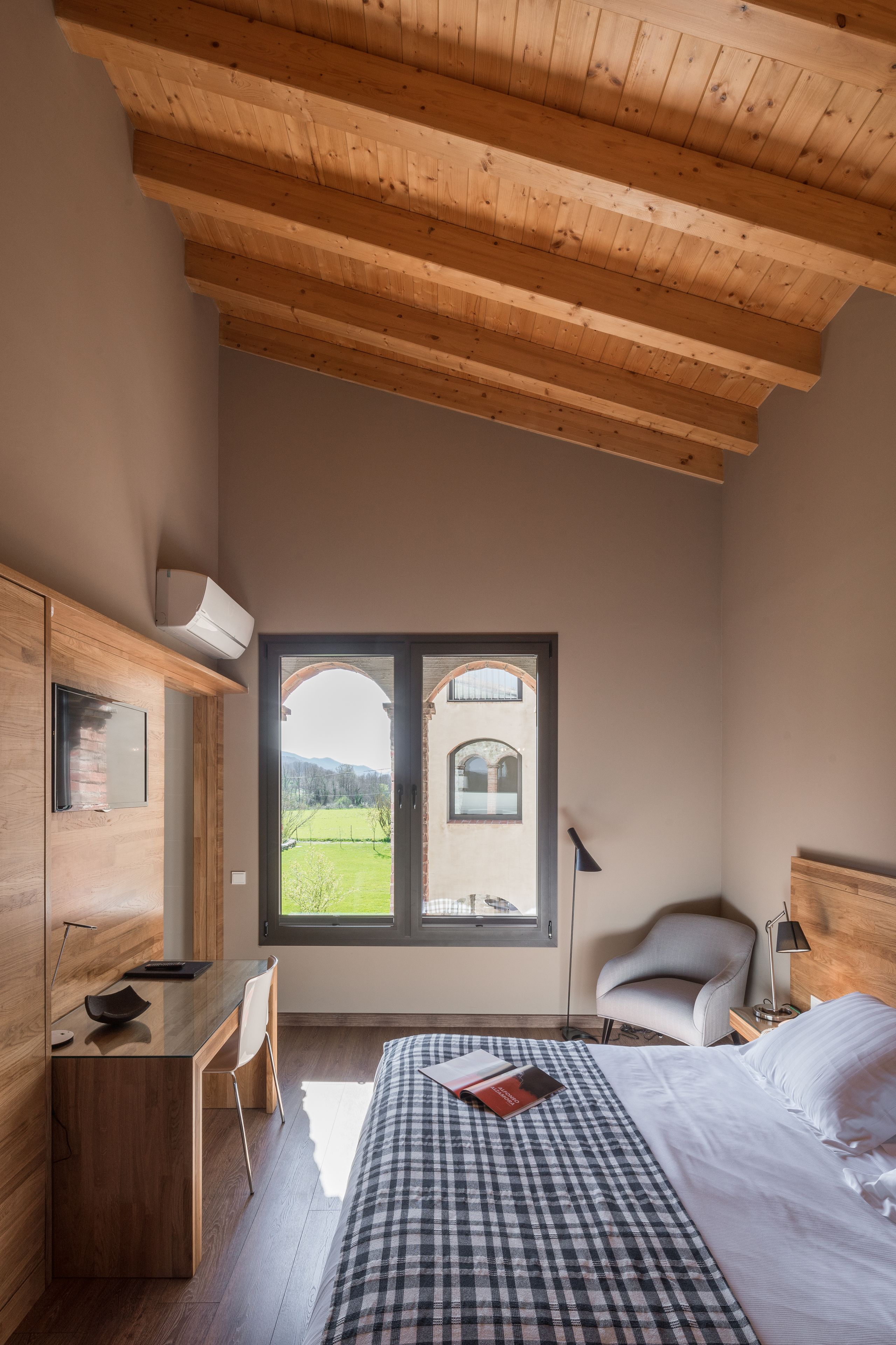 Un dormitorio con techo de madera, un escritorio, una silla, una cama con colcha a cuadros y vistas a un paisaje verde por la ventana.