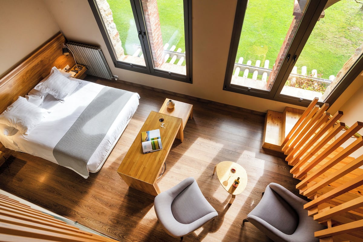 A bedroom with a bed, a wooden table, two chairs, and a view of a grassy yard through large windows.