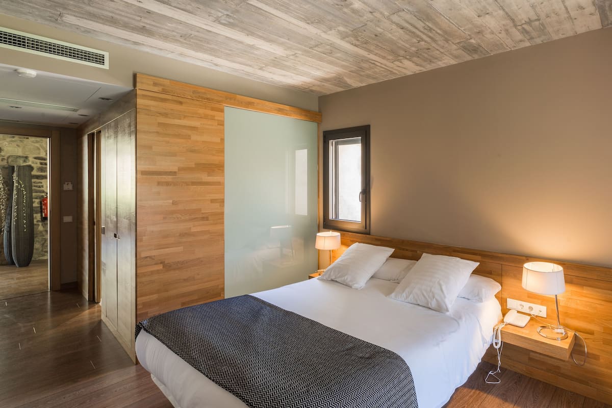 A hotel room with a wooden headboard, a large bed with white linens, two bedside lamps, and a wooden nightstand.