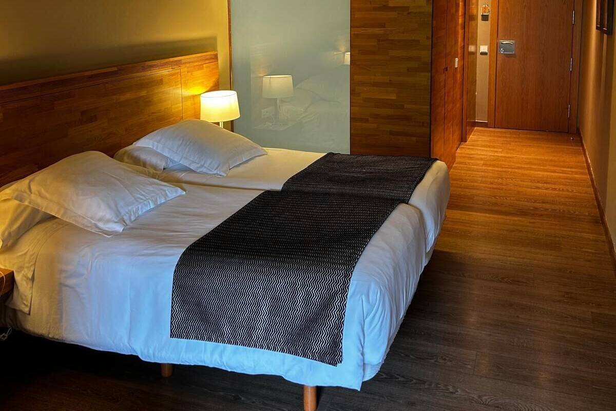 A hotel room with a wooden ceiling, a bed with white bedding, a patterned throw blanket, and a nightstand with a lamp.