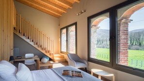 A modern hotel room with a wooden staircase, a bed with a magazine, a desk with a lamp, and large windows offering a view of the countryside. - Les Planes del Grau (Sant Joan de Les Abadesses)