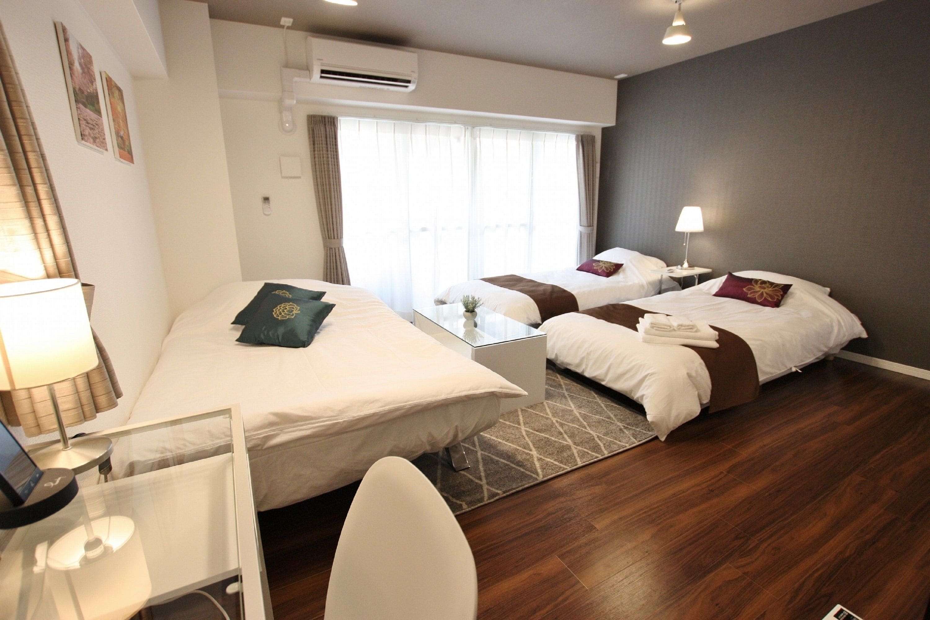 A hotel room with two beds, a desk, and a chair. There is a window with curtains, an air conditioning unit, and a lamp on a bedside table.