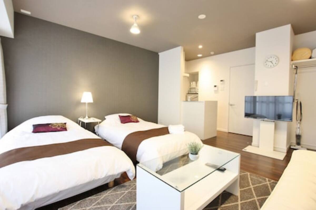 Habitación de hotel con dos camas, un pequeño rincón de descanso, un televisor, un reloj y una kitchenette al fondo.