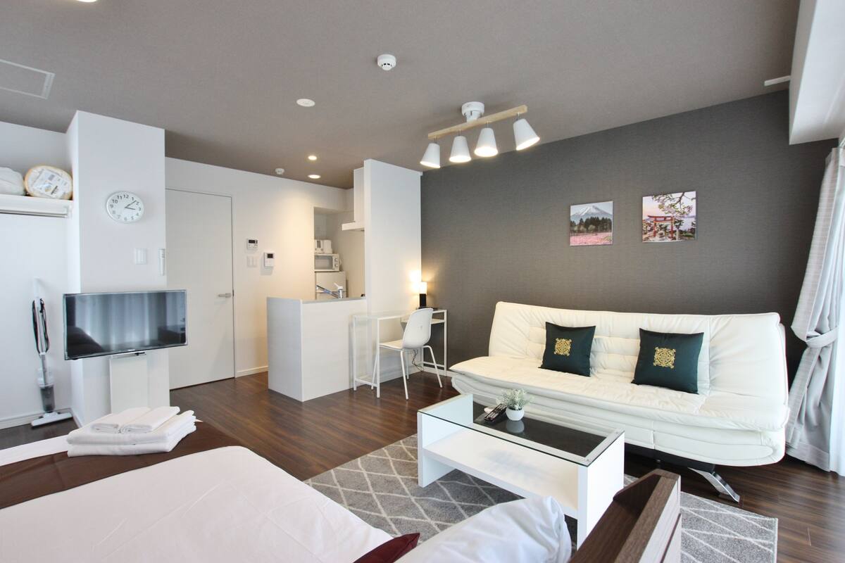 Una habitación de hotel moderna con una cama, un sofá blanco, una mesa de centro, un televisor y una pequeña cocina.