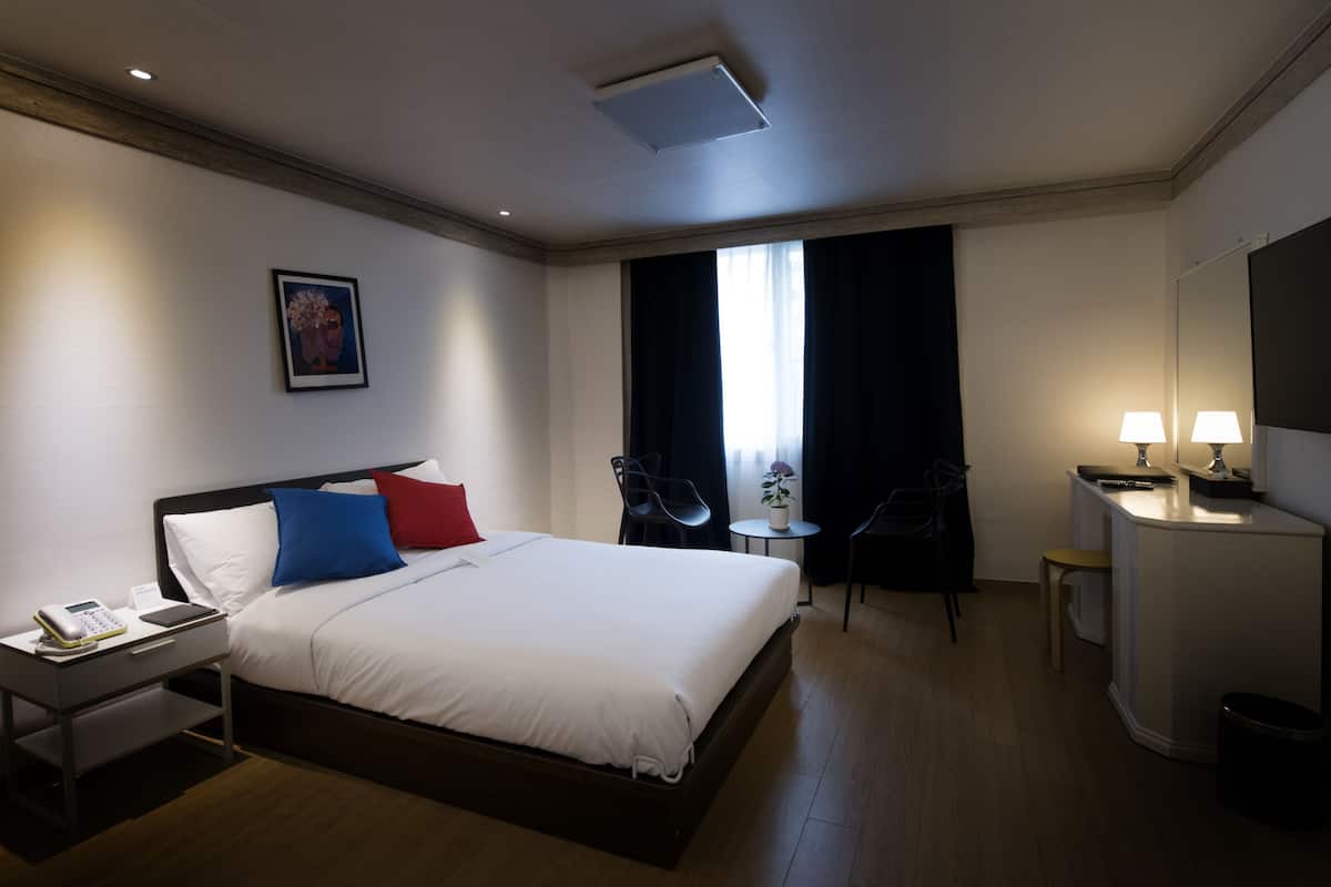 Habitación de hotel con una cama grande, mesita de noche con teléfono, pequeño escritorio con lámpara, una silla, ventana con cortinas y un cuadro en la pared.