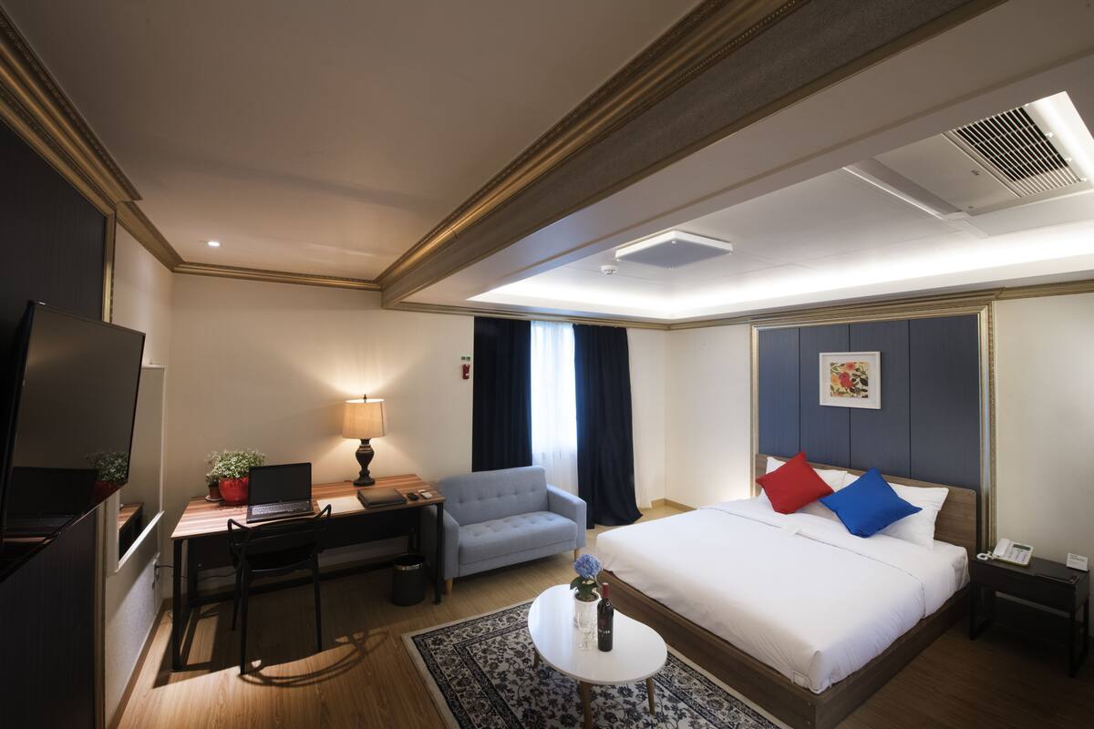Una habitación de hotel con cama, escritorio, silla, televisión y una mesita con una botella y una flor.