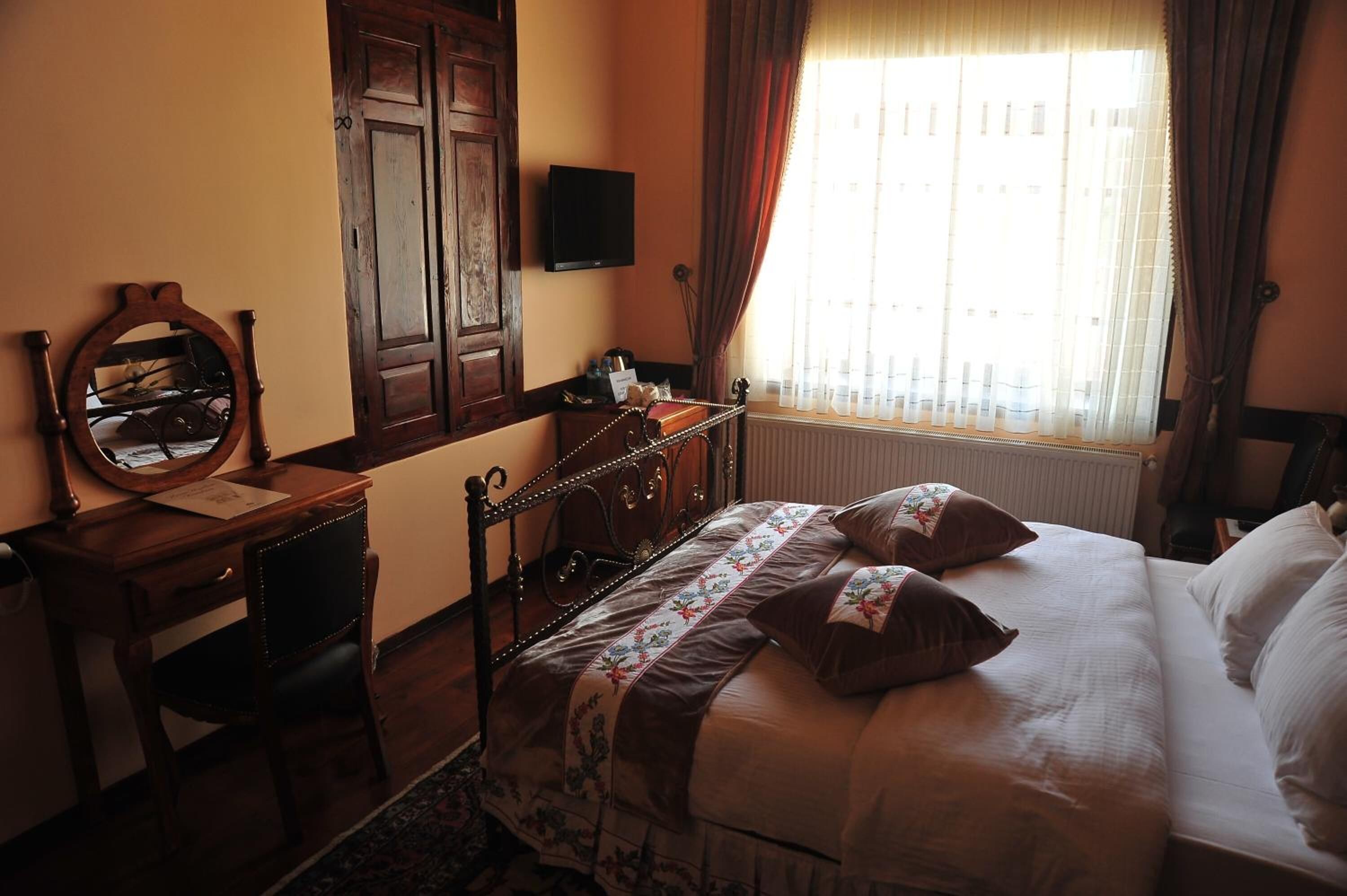A bedroom with a bed, a desk, a chair, a mirror, a television, and a window with curtains.
