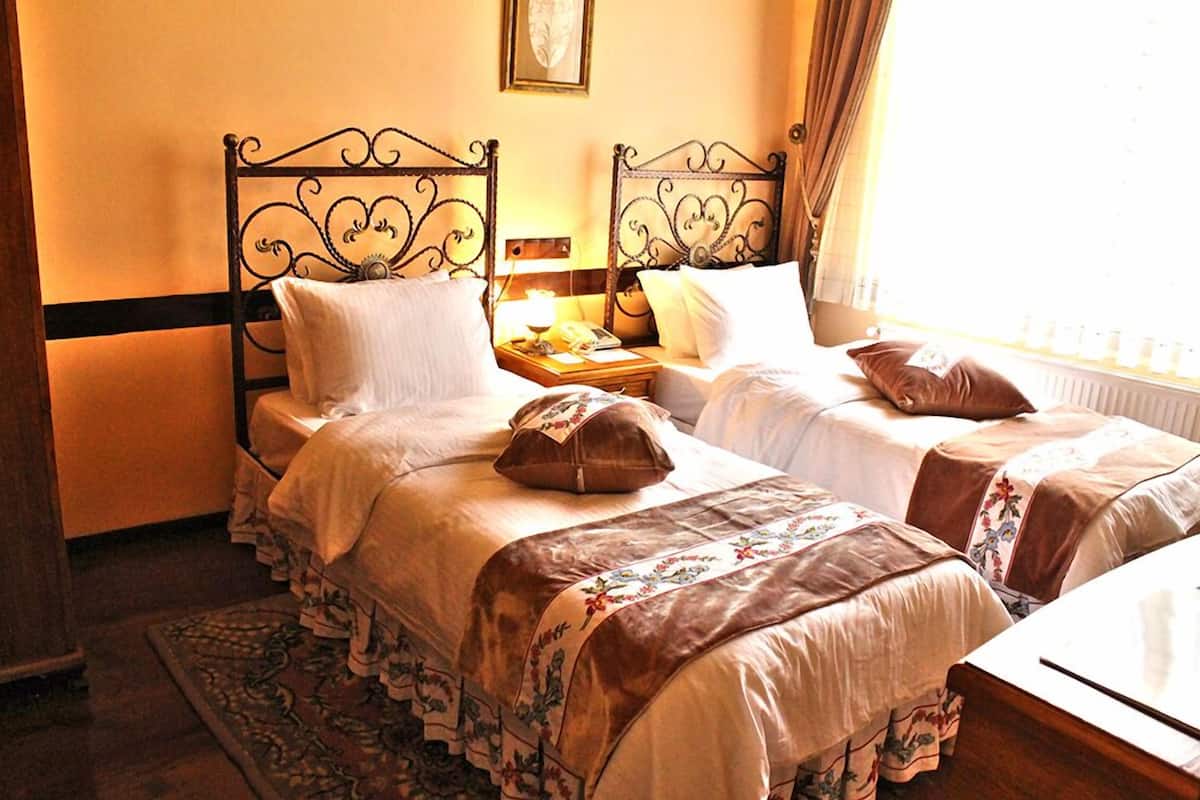 A room with two beds, a nightstand with a lamp, a wooden headboard, a framed picture on the wall, and a window with curtains.