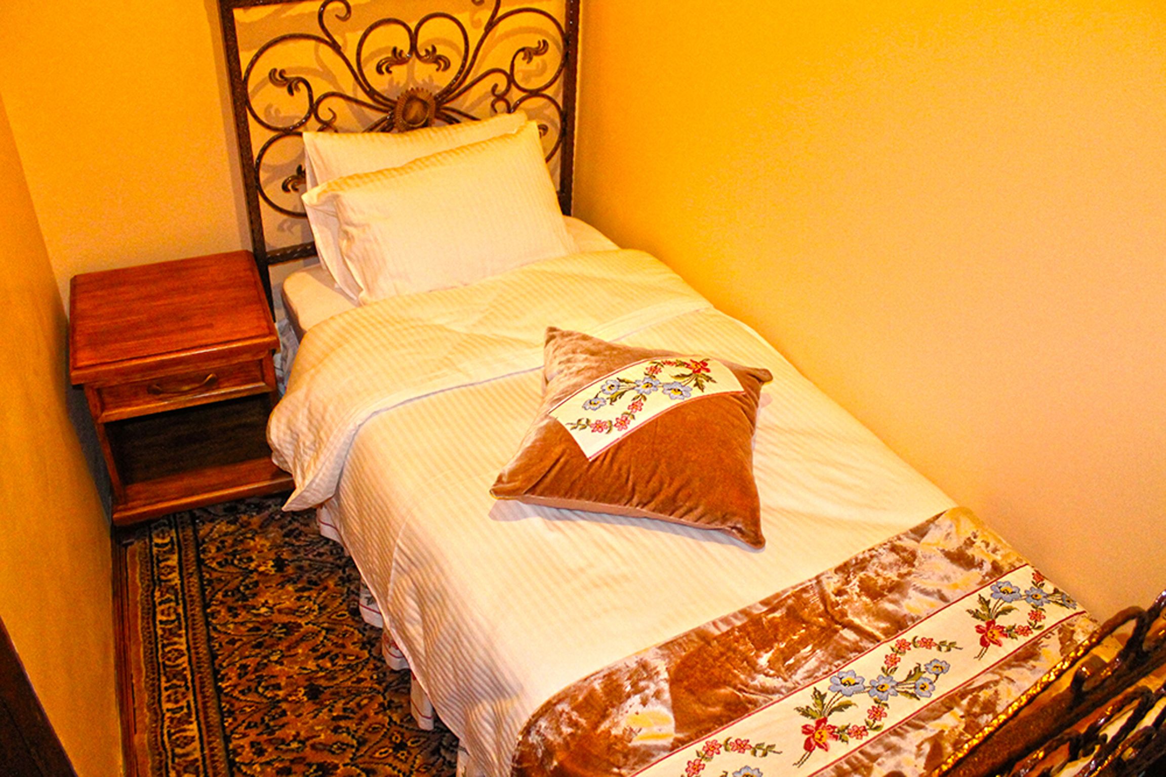 A neatly made bed with a floral-patterned bedspread, a wooden nightstand, and a patterned rug.