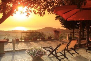Una terraza con vistas a la puesta de sol sobre el mar, plantas en macetas y un paraguas rojo.