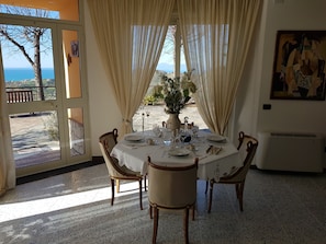 Un coin repas avec une table dressée pour six personnes, un vase de fleurs et une vue sur la mer par de grandes fenêtres.