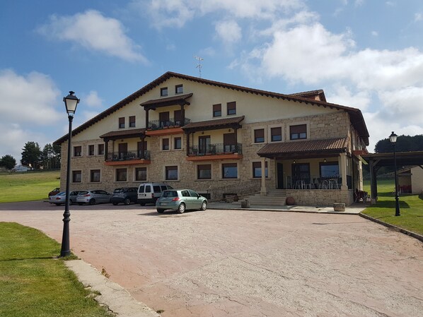 Un edificio de varios pisos con fachada de piedra, varios balcones y un camino pavimentado con coches estacionados.
