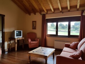 Una sala de estar con techo de madera, un televisor sobre una mesa de madera, un sillón rojo, un sofá color beige y una ventana con vistas a la vegetación.