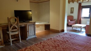 Una sala de estar con un escritorio de madera, televisión, alfombra estampada, una silla y una ventana con cortinas.