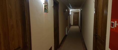 Un pasillo largo y estrecho con puertas de madera a ambos lados, un radiador a la izquierda y un tablón de anuncios rojo a la derecha.