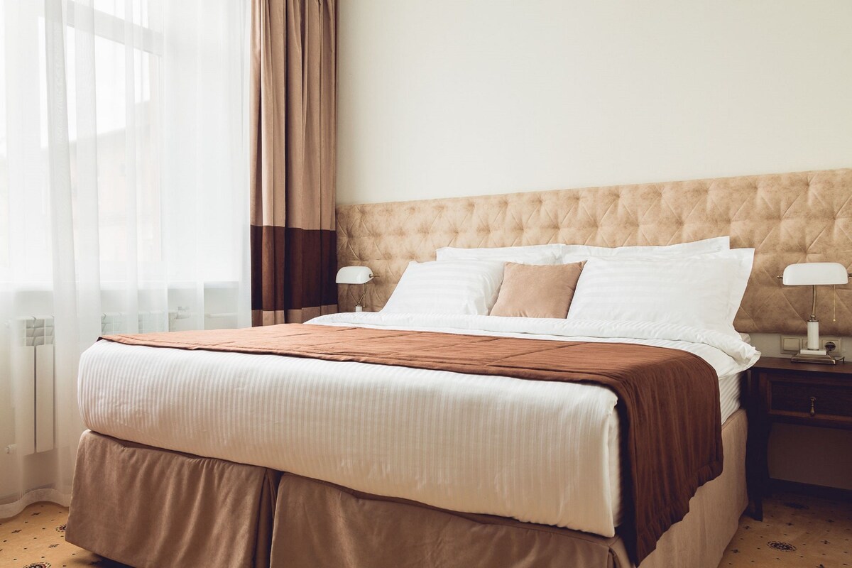 A hotel room with a large bed, a headboard, two bedside tables with lamps, and a window with curtains.