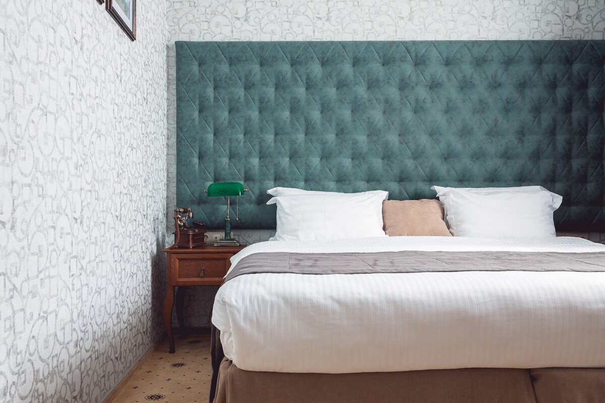 A neatly made bed with white and beige bedding, a green headboard, and a small wooden nightstand with a green lamp.
