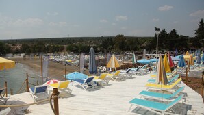 A beachside area with rows of sun loungers and umbrellas, a wooden deck, and a view of the beach and distant landscape. - Queenaba Beach (Erdemli)