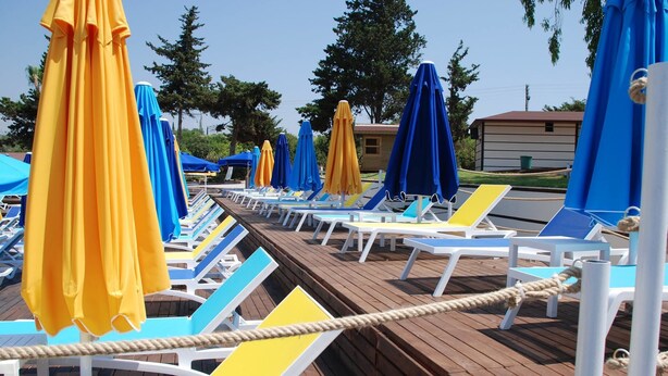 A deck with rows of sun loungers and umbrellas, a rope barrier, and a small building in the background. - Queenaba Beach (Erdemli)
