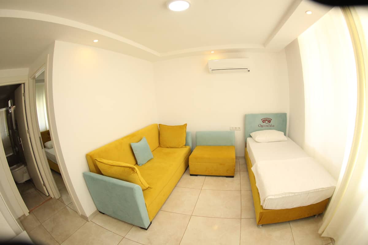 A compact room with a yellow sofa, a matching ottoman, and a single bed with a white mattress.