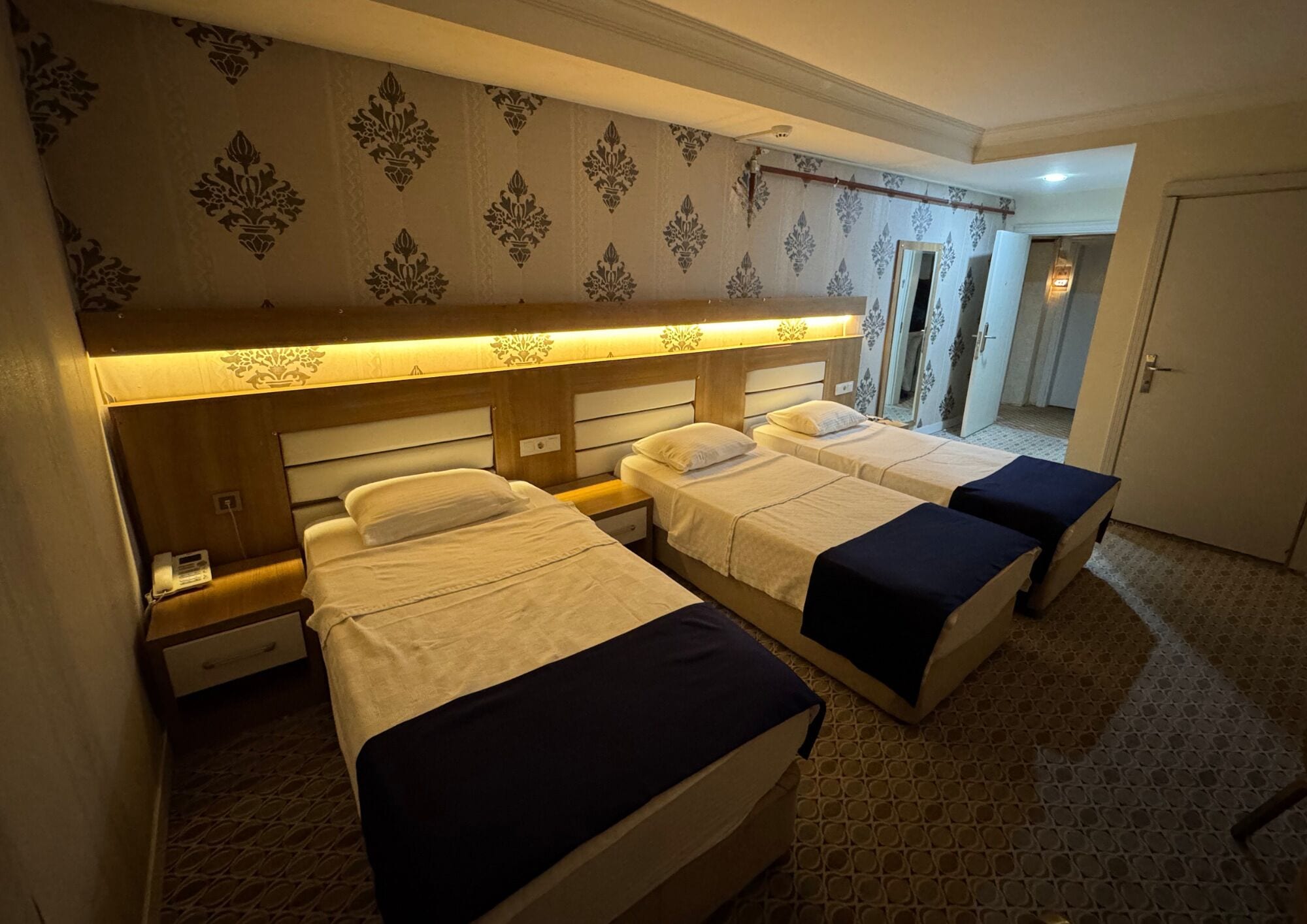 A hotel room with three beds, a wooden headboard, a patterned wallpaper, and a tiled floor.