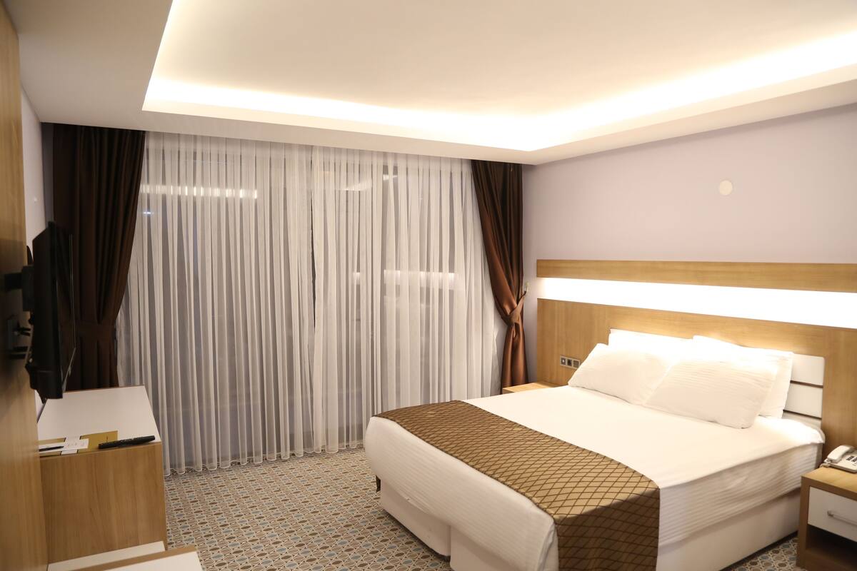 A hotel room with a bed, bedside table, lamp, curtains, and a TV.