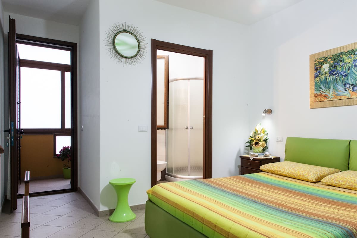 A bedroom with a bed, a green nightstand, a green stool, a door, and a mirror on the wall.