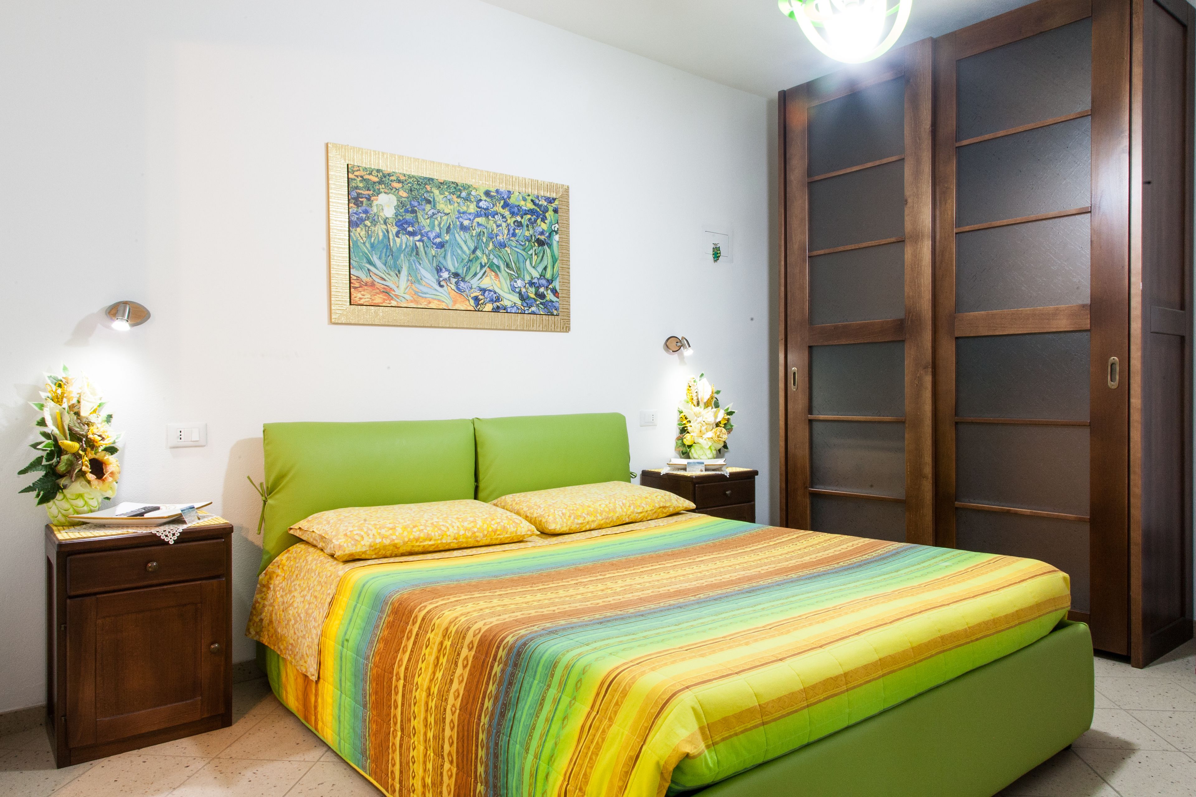 Un dormitorio con una cama verde, una colcha a rayas de colores, dos mesitas de noche con lámparas, un armario de madera y un cuadro con motivos florales en la pared.