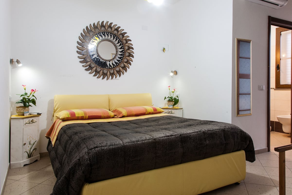 A bedroom with a bed, a bedside table with a lamp, a mirror, and a door leading to another room.