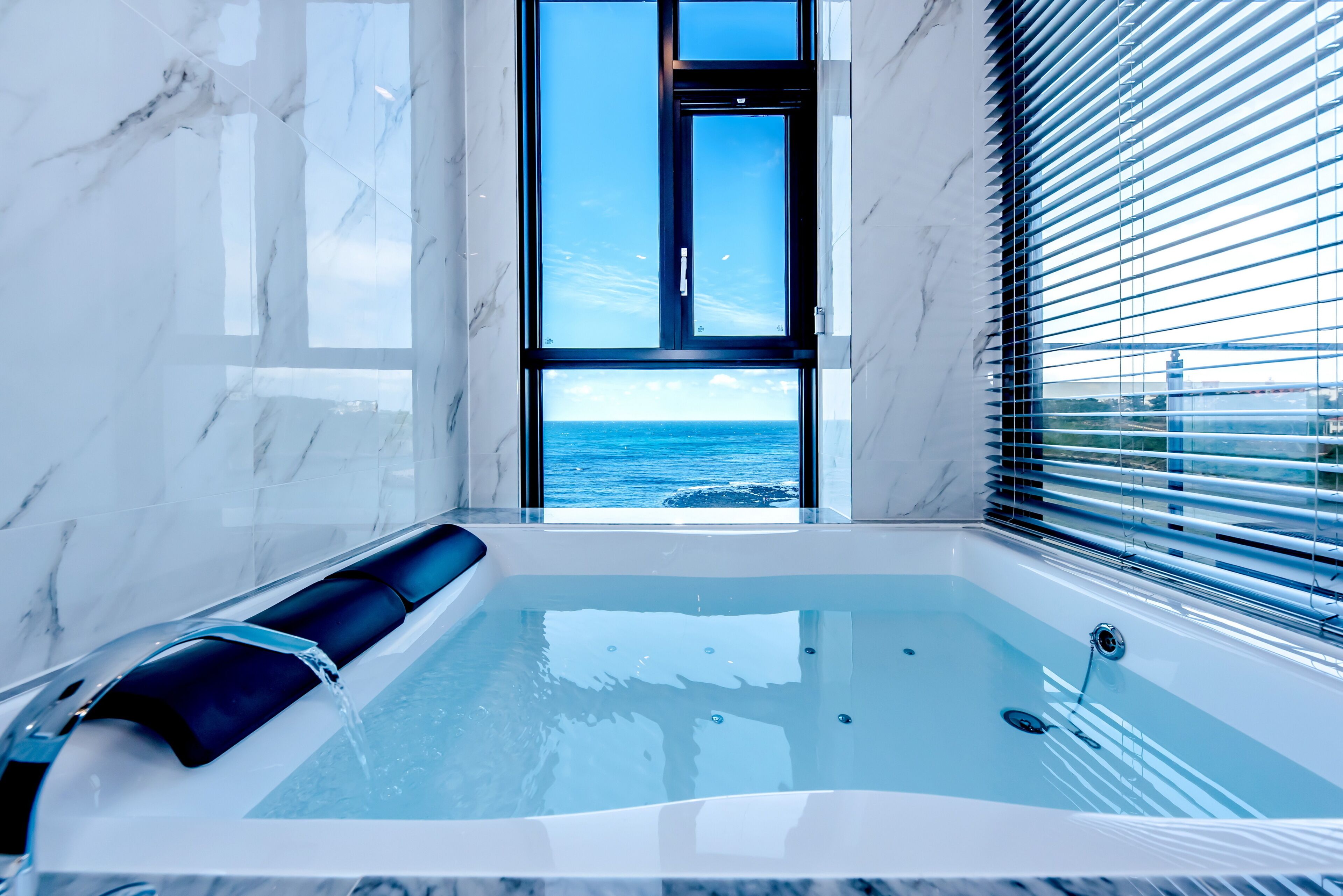 A modern bathroom with a marble wall, a bathtub with a black seat, and a view of the ocean through a window.