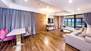 Une chambre d’hôtel moderne avec un plancher en bois, une table à manger avec des chaises roses, un canapé, une télévision et de grandes fenêtres.
