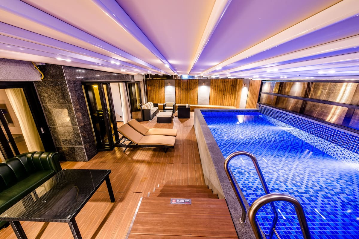 Un espace piscine intérieur moderne, doté d’une paroi en verre, de chaises longues et d’un plancher en bois.