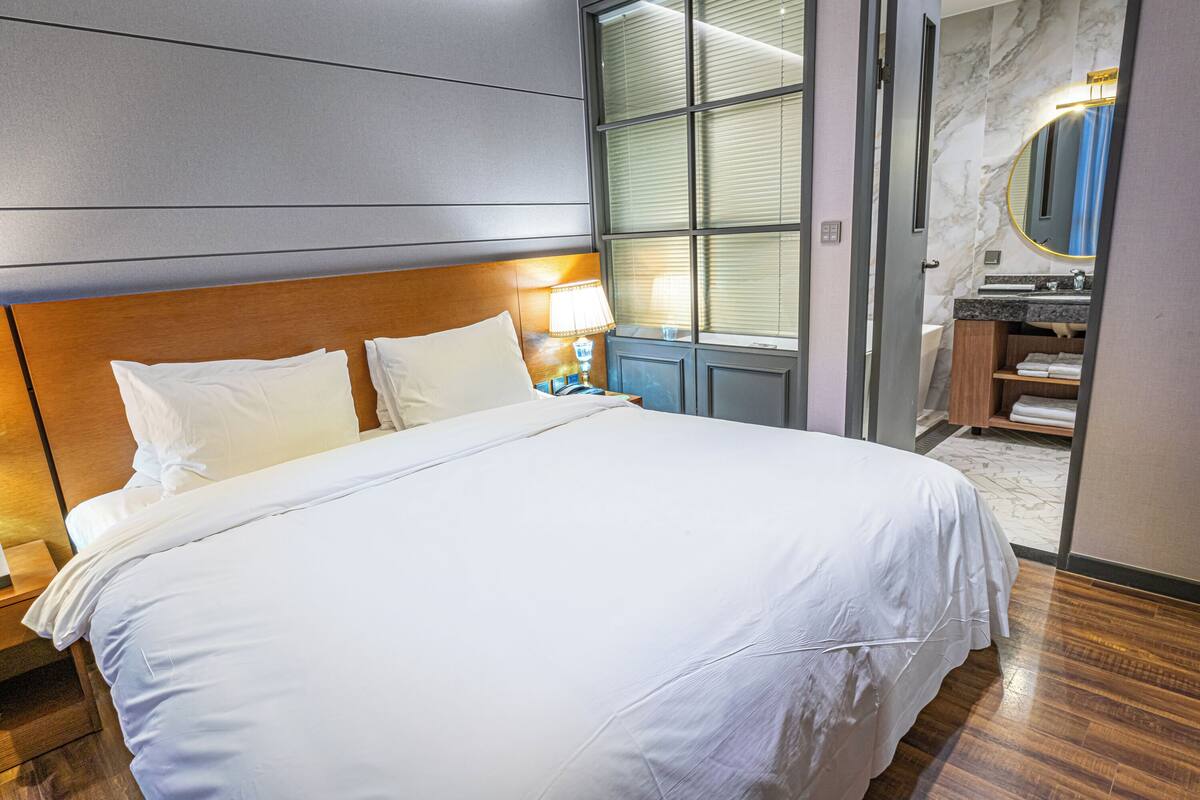 Une chambre d’hôtel avec un grand lit, une table de chevet, une lampe, une salle de bain avec un lavabo, un miroir et un porte-serviettes.