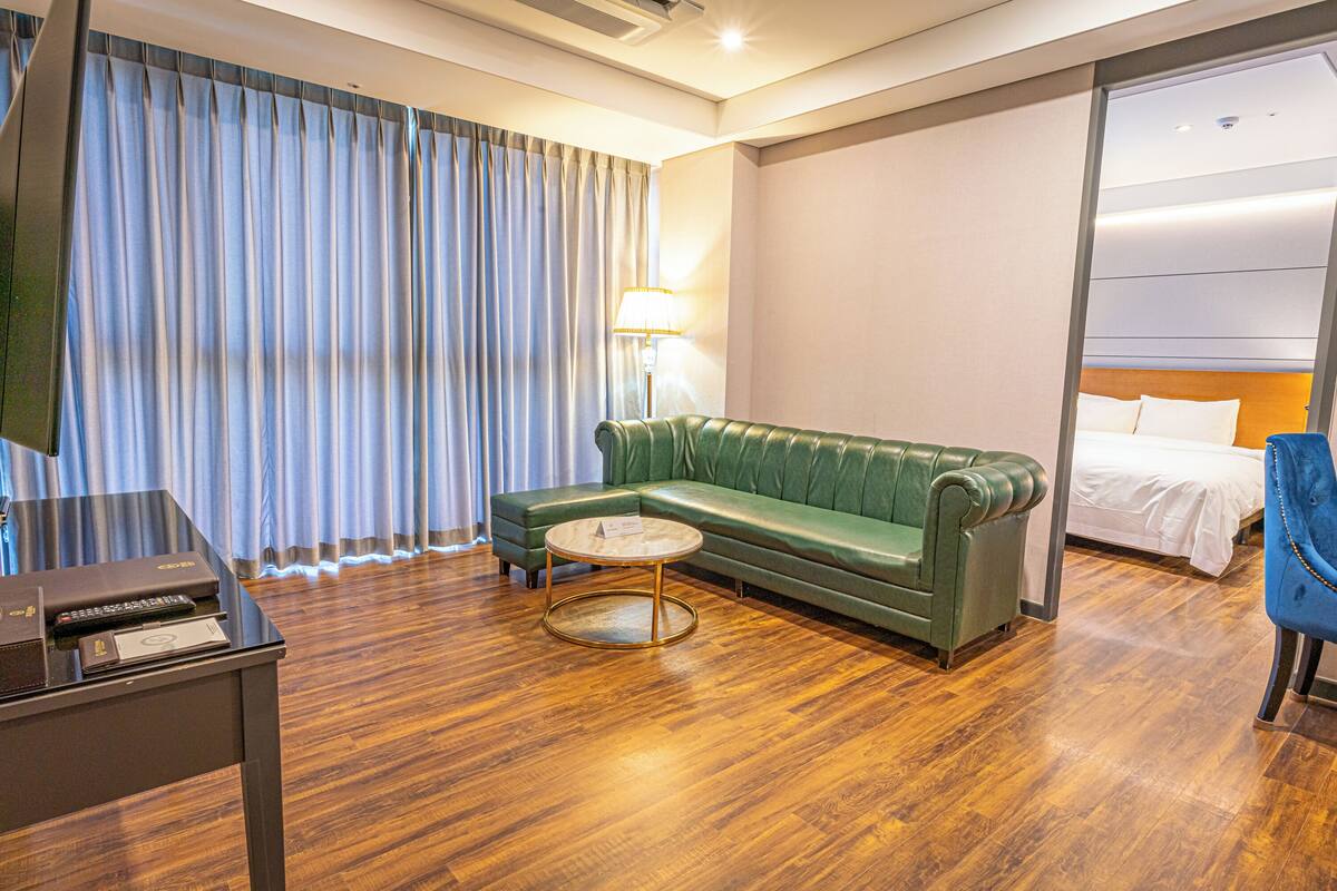 Une chambre d’hôtel avec un canapé vert, une table basse ronde, un lit avec du linge de maison blanc, une chaise bleue et un téléviseur à écran plat.