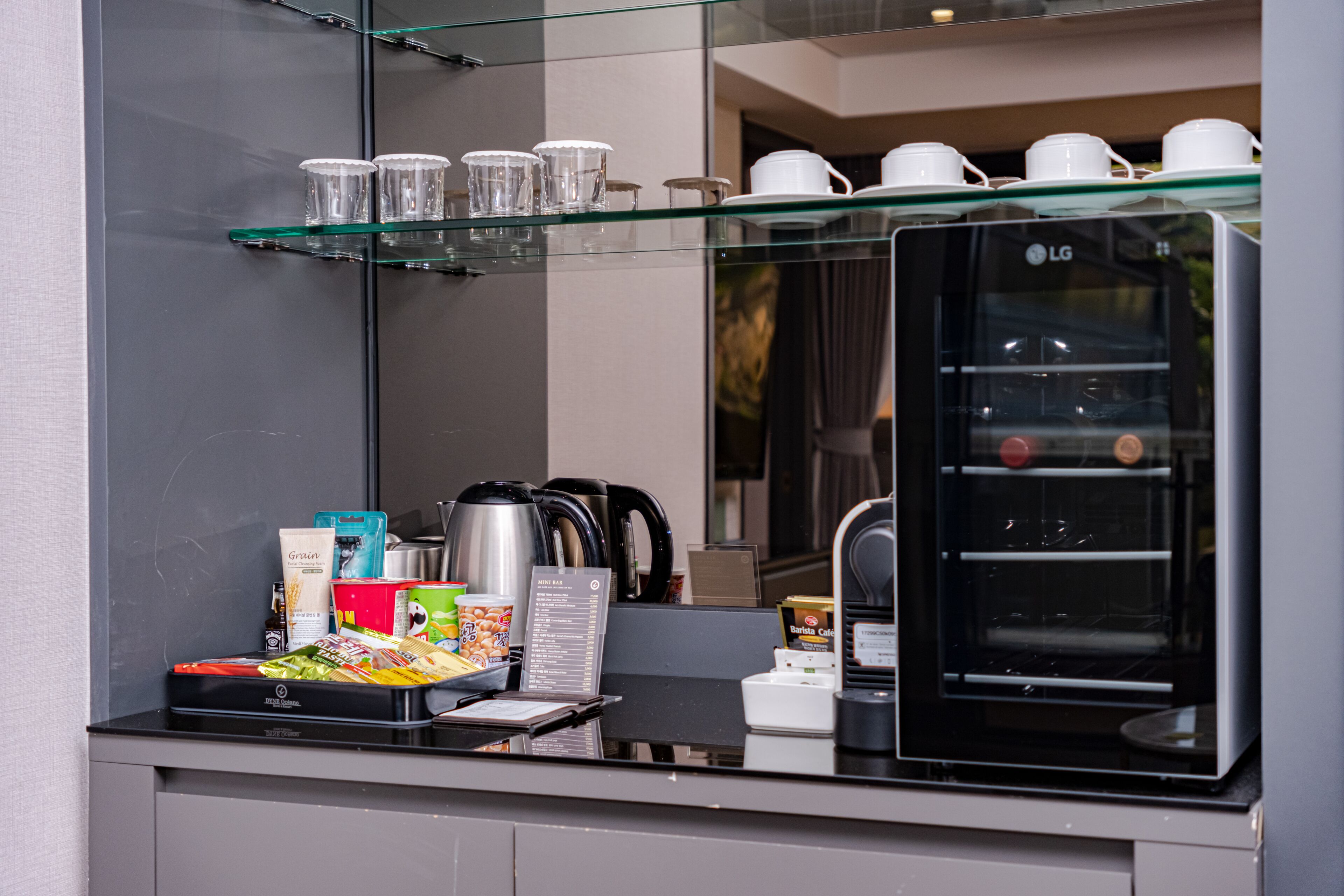 Un espace cuisine moderne équipé d’une machine à café, d’une bouilloire et d’un four à micro-ondes. Des étagères en verre, situées au-dessus du plan de travail, servent à ranger des tasses et une cafetière.