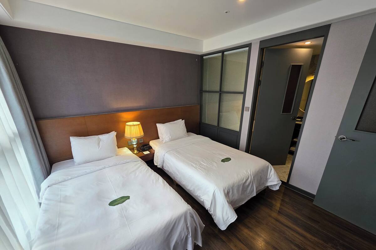 Une chambre d’hôtel avec deux lits, une table de chevet avec une lampe, une tête de lit en bois et une porte donnant sur une autre pièce.