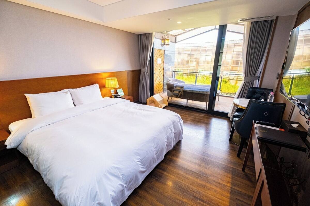 Une chambre d’hôtel avec un grand lit, un bureau et une chaise. Il y a une fenêtre avec des rideaux et une vue sur la verdure à l’extérieur.