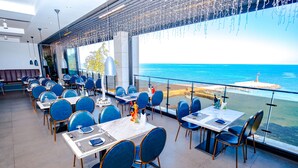 A restaurant with tables and chairs arranged for dining, large windows offering a view of the sea, and a modern interior design.