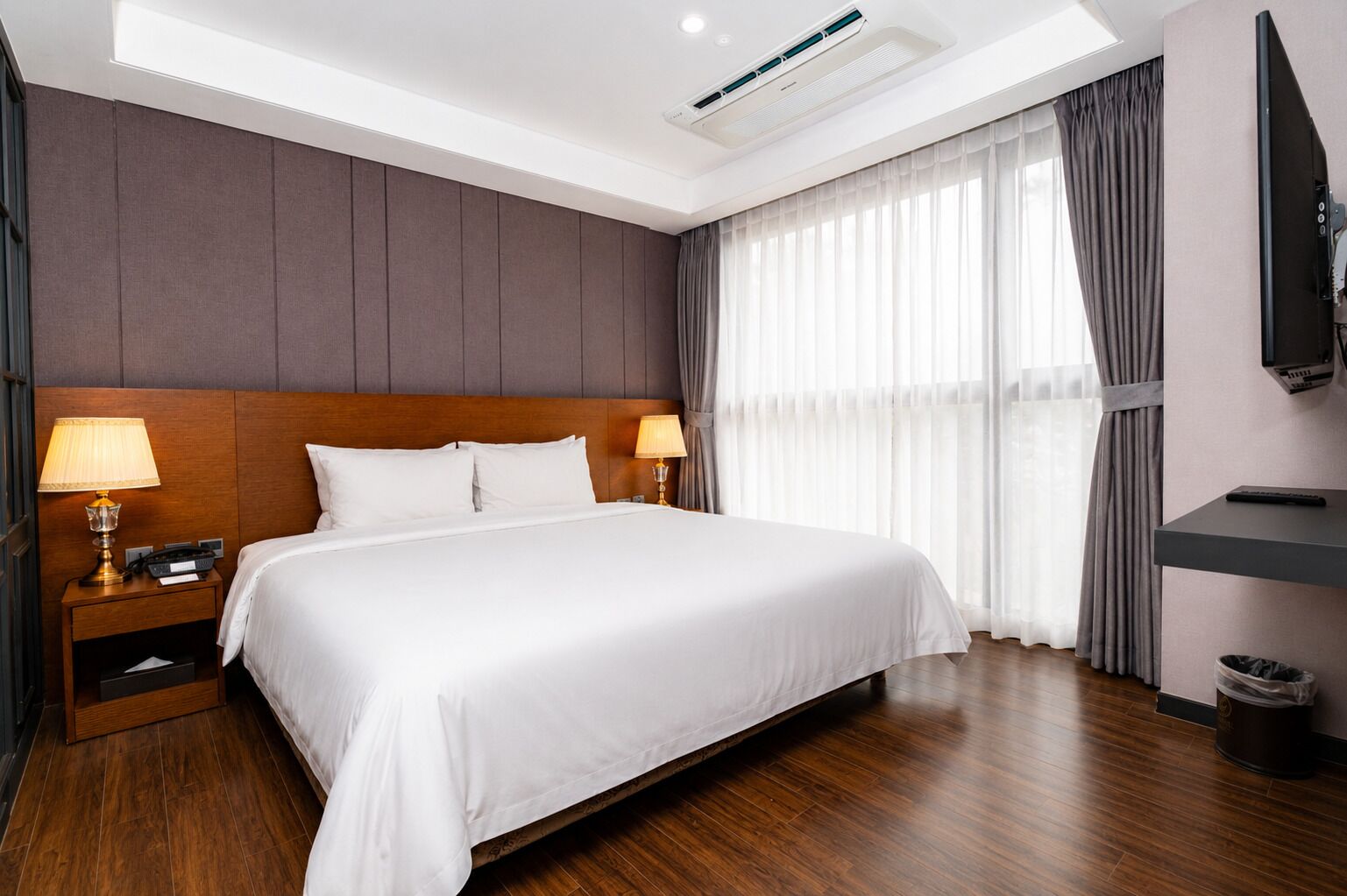 A hotel room with a large bed, two bedside lamps, a flat-screen TV, and a ceiling-mounted air conditioner.
