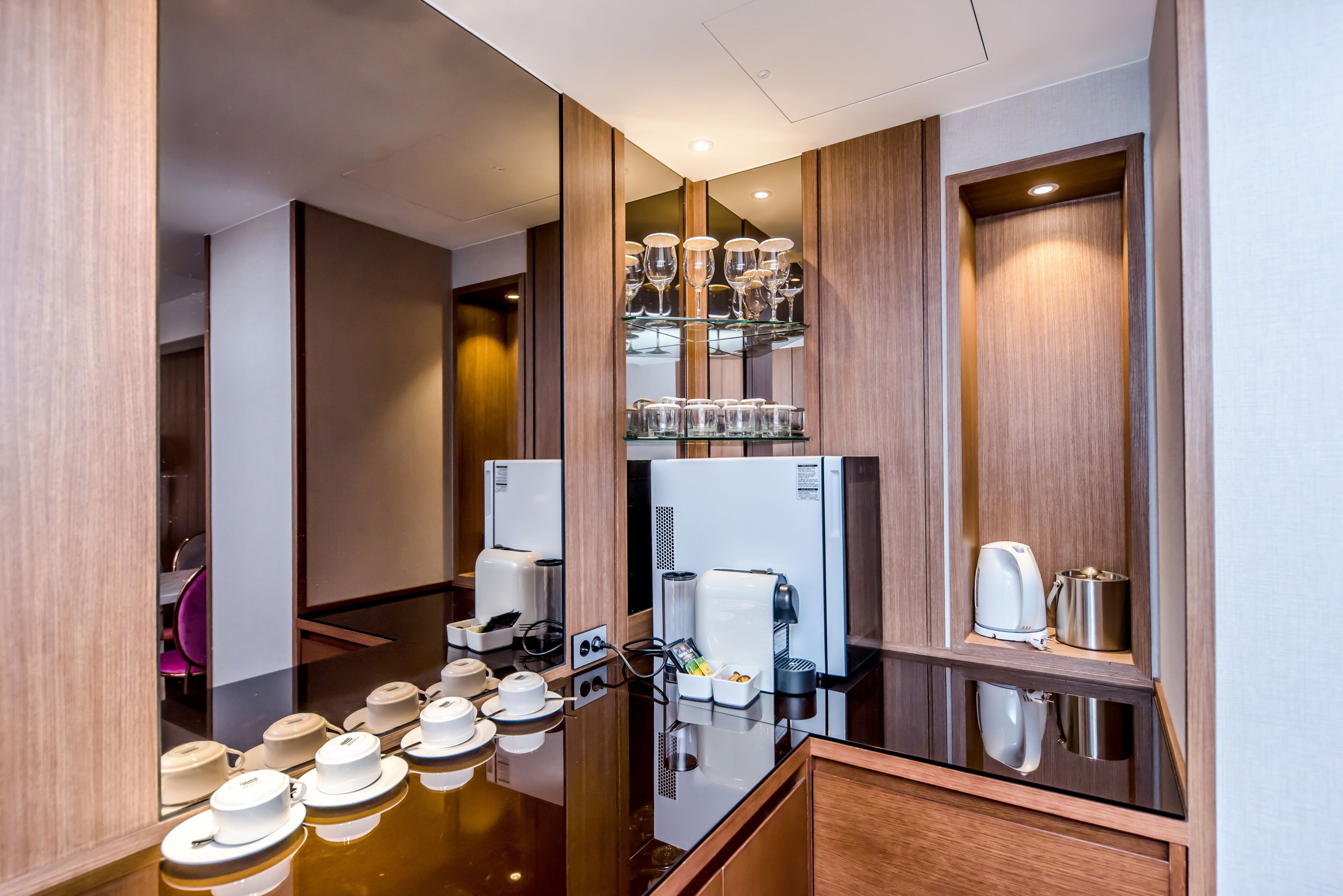 A modern hotel room with a wooden panel design, a glass-top dining table with tea sets, a coffee machine, and a refrigerator.