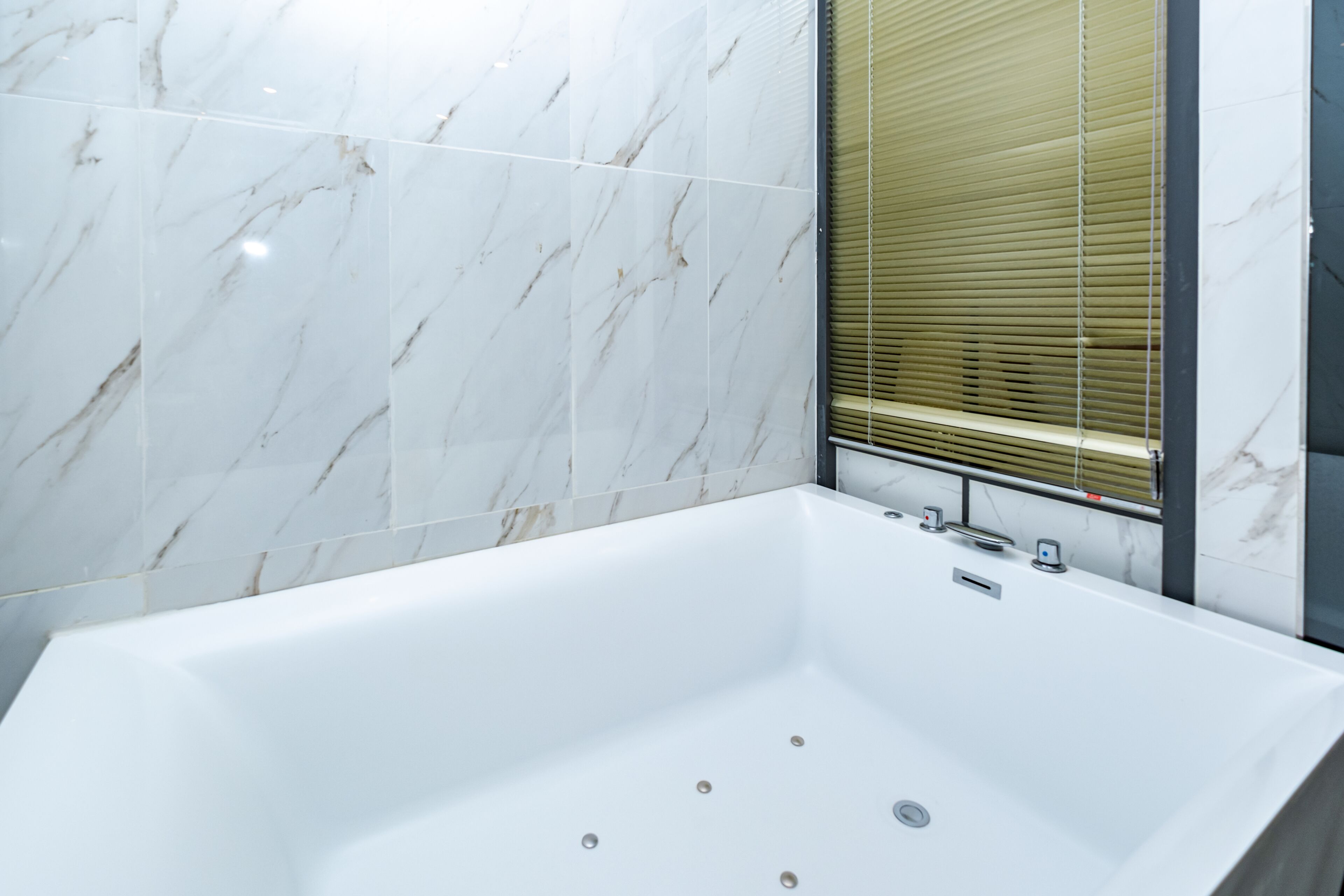 A modern bathroom with a large white bathtub, marble walls, and a window with blinds.